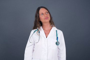 Pleasant looking young doctor female student, looks pensively aside, plans actions after work, imagines what to do. Clever woman thinks over about new project. Copy space on gray.