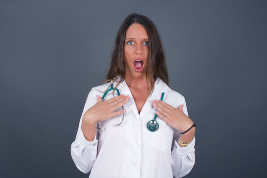 Shocked scared beautiful young doctor woman keeps hands on chest, mouth widely opened, stares at camera realizes that she forgot to pay bills isolated indoors. Girl saying: Who, me?