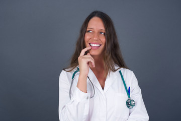 Dreamy doctor female with thoughtful expression, looks to the camera, keeps hand near face, bitting a finger thinks about something pleasant, poses against gray wall.