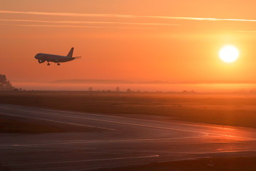Fototapeta na wymiar The photo shows a plane landing at dawn or sunset. The crimson rays of the sun nicely illuminate the runway. The photo was taken on a summer fog day at the airport in the Siberian city of Novosibirsk.