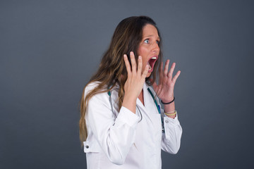 Portrait of young doctor woman with shocked facial expression holding hands near face, screaming and looking sideways at something amazing. Stands against gray studio Wall.