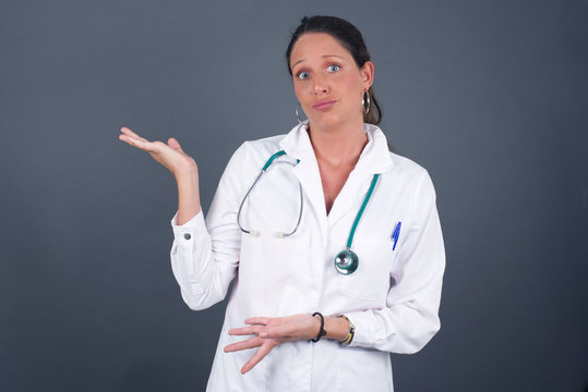 Beautiful European doctor girl pointing aside with both hands showing something strange and saying: I don't know what is this. Standing against gray background. Advertisement concept.