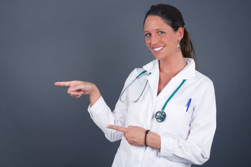 Positive attractive young doctor female, points aside with cheerful and surprised expression with mouth opened, shows something amazing standing indoors. Advertisement concept.