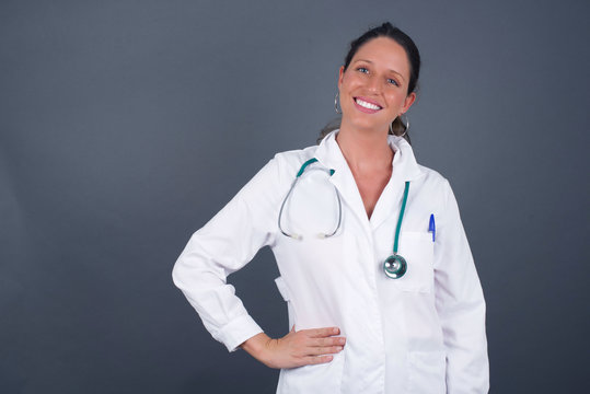 Studio shot of cheerful doctor woman keeps hand on hip, smiles broadly, anticipates for surprise prepared by friends on her birthday, isolated over gray background.
