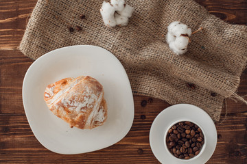 cup of coffee, croissant, burlap, coffee beans and cotton on brown wooden table