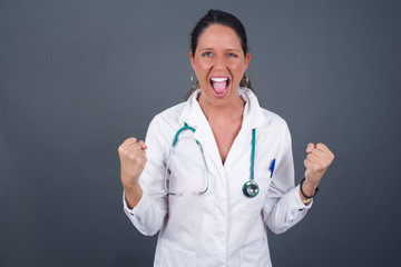 Portrait of beautiful doctor female looks with excitement at camera, keeps hands raised over head, notices something unexpected, isolated over gray wall. Lovely woman reacts on sudden news.