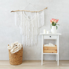 Spring boho home interior decor: macrame wall hanging decoration, white bedside table, ceramic vase with bouquet of pink tulips flowers, candles, wicker basket, plaid. Light cozy modern stylish room.