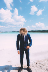 Portrait of blond curly man dressed blue business suit. Clear water and blue sky on the background. Summer time. Travel topic.