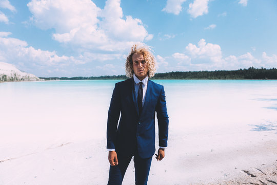 Portrait Of Blond Curly Man Dressed Blue Business Suit. Clear Water And Blue Sky On The Background. Summer Time. Travel Topic.