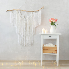 Spring boho home interior decor: macrame wall hanging decoration, white bedside table, vase, bouquet of pink tulips flowers, lighted candles, led garland glowing lights. Light cozy modern stylish room © NesolenayaAleksandra