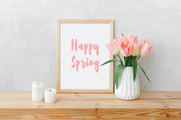 Frame with text HAPPY SPRING, white ceramic vase with bouquet of pink tulips flowers, candles on a wooden table or shelf on a background of light gray wall. Stylish spring home interior decor. Mock up