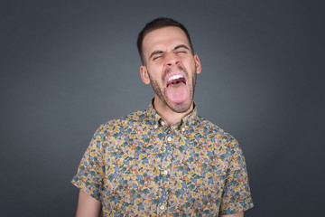 Beautiful caucasian man with happy and funny face smiling and showing tongue. Wearing casual clothes and standing against gray studio background.