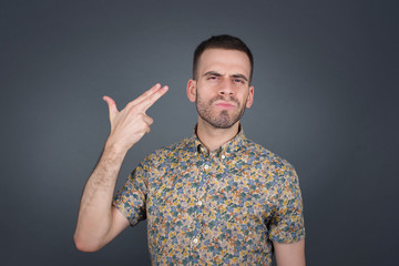 Fototapeta premium Unhappy blond young male imitates gun shoot makes suicide gesture keeps two fingers on temples has puzzled expression dressed in casual clothes stands indoors.