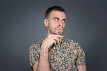 Dreamy male with pleasant expression, wearing casual clothes, looks sideways, keeps hand under chin, thinks about something pleasant, poses against gray background.