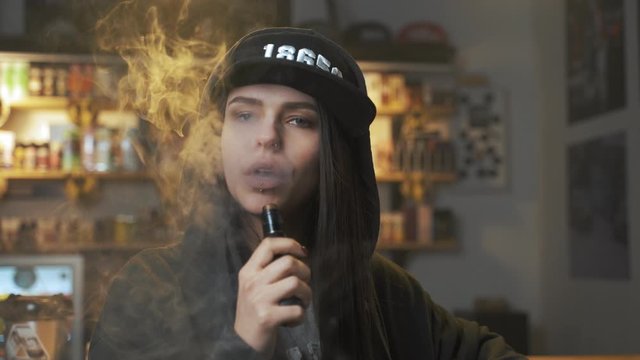 Young pretty woman in cap smoke an electronic cigarette at the vape shop. Hip-hop style. Closeup.