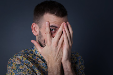 Anxiety - a conceptual image of a beautiful young caucasian man covering his face with his hands and peering out with one eye between her fingers standing indoors. Scared from something or someone.