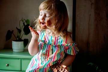 Cute curly haired baby girl eating chocolate,child smeared. dirty cheeks, nose and hands. Chocolate celebration on July 7th. harmful sweets for children. unhealthy food. indoor plants.