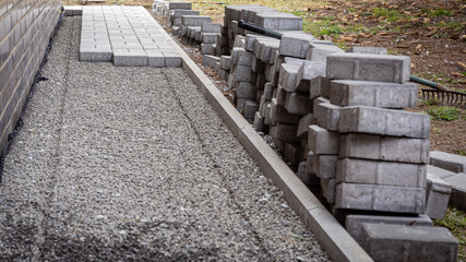 Laying gray concrete paving slabs in a house walkway. Concrete blocks and a curb are professionally installed on leveled gravel foundation base. Stack of concrete blocks ready for laying