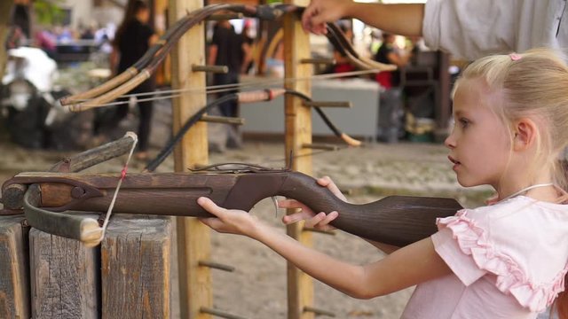 Blond child girl aims and shoots with crossbow at open air amusement park medium shot
