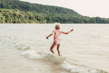 Cute funny Caucasian girl walking in lake river waves at sunset. Child jumping water on a beach. Authentic real lifestyle happy childhood. Summer fun outdoor aquatic activity. View from back.