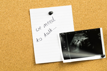We need to talk text  message on white notebook paper card with an ultrasound photo of baby fetus - proof of pregnancy. Pregnancy announcement on cork board.