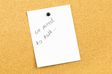 We need to talk message written on white card of paper notebook, hanging on cork card board. Communication problems in family or work office space.