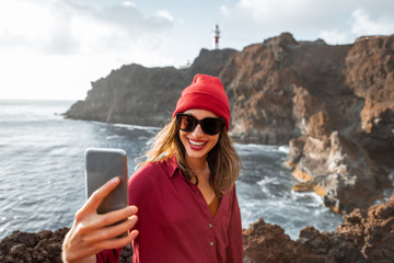 Young stylish woman making selfie portrait while traveling on the rocky ocean coast near the lighthouse on Tenerife island, Spain - Powered by Adobe