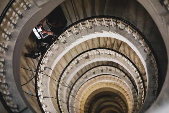 Shallow depth of field (selective focus) image with a woman journalist using her laptop and mobile phone on a very deep spiral staircase