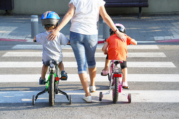 Mother goes pedestrian crossing with children on bicycles. Mother with son and daughter crossing the road in the city. Back view.