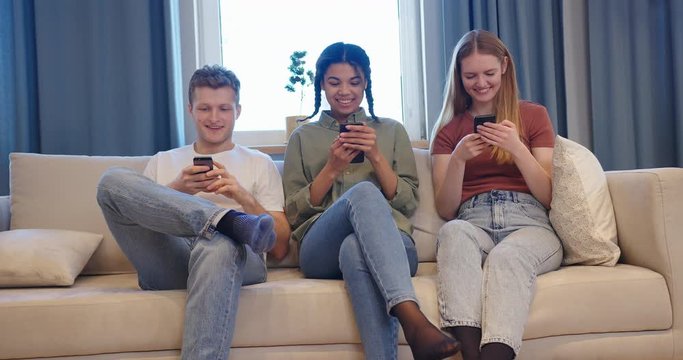 Happy young multiethnic group of friends sitting on the sofa and watching funny pictures on their smartphones
