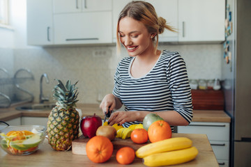 Happy healthy woman cutting fruits on a wooden board while making breakfast in a kitchen