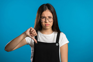 Young asian girl standing on blue studio background expressing discontent and showing thumb down...