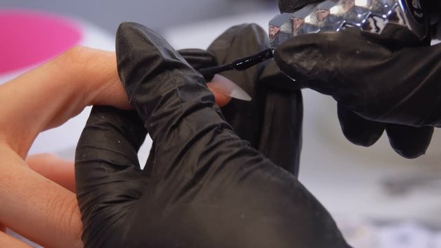 Close up of a manicurists hands in black gloves painting manicured nails with a black gel 