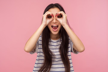 Portrait of overjoyed surprised girl with long brunette hair looking through binoculars shaped with fingers, zooming at camera and expressing shock, amazement. studio shot isolated on pink background