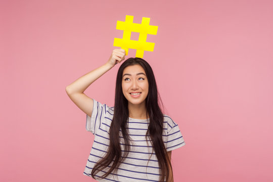 Portrait of positive trendy girl with brunette hair in striped t-shirt holding hashtag symbol on head and smiling, tagging famous blog, viral content. indoor studio shot isolated on pink background