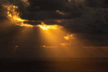 Long view of sun cast its golden rays down upon sea surface through dark dramatic clouds
