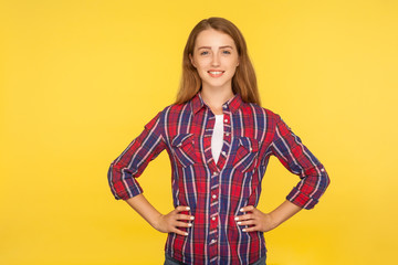 Portrait of happy beautiful ginger girl with charming smile and in checkered shirt holding hands on hips, looking at camera with confident expression. indoor studio shot isolated on yellow background