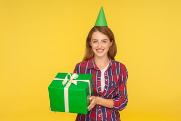 Portrait of cheerful funny ginger girl with party cone on head holding gift box and smiling happily, excited about present on birthday, holiday bonus. indoor studio shot isolated on yellow background