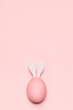 Huevo de pascua de color rosa pastel con orejas de conejo de color blanco sobre fondo rosa pastel vertical.