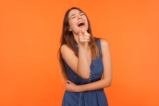 Lol, hey you! Excited joyful brunette woman laughing out loud, holding her belly and pointing finger to camera, teasing mocking, making fun of people. indoor studio shot isolated on orange background