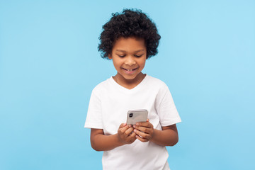Happy adorable excited little boy with curly hair reading funny message on smartphone and smiling, satisfied with children mobile application, using cellphone. studio shot isolated on blue background © khosrork