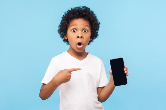 Wow awesome mobile app. Amazed excited cute little boy pointing at cell phone and looking at camera with shocked face, child surprised by telephone, online service. indoor studio shot blue background