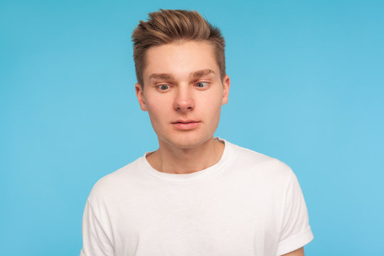 Portrait of funny comic young man in casual white t-shirt looking cross-eyed with awkward silly dumb expression, having fun alone, playing fool. indoor studio shot isolated on blue background, closeup