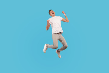 Fototapeta na wymiar Full length, excited successful man in white t-shirt and casual pants jumping with happiness and shouting yes, I did it, feeling inspired and energetic. indoor studio shot isolated on blue background