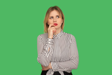 Beautiful pensive girl with red lips in striped blouse standing with thoughtful serious smart expression, pondering answer, having doubts and suspicion. indoor studio shot isolated on green background