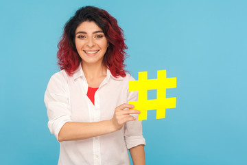 Blogs and popular topics. Charming happy woman with fancy red hair holding big hashtag symbol and smiling to camera, hash sign to tag forum discussions. indoor studio shot isolated on blue background - obrazy, fototapety, plakaty