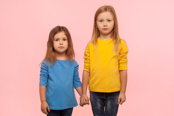 Little friends. Two cute charming preschool girls in sweatshirts standing together, holding hands and looking at camera with serious calm expression. indoor studio shot isolated on pink background