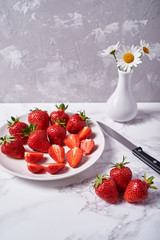 Red ripe strawberries in white ceramic bowl and chamomile flowers in vase on grey background, copy space. Healthy food concept, still life