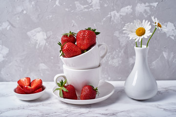 Ripe organic strawberries in white ceramic bowl and chamomile in vase on gray concrete background, copy space. Healthy food concept, still life