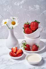 Ripe organic strawberries in white ceramic bowl and chamomile in vase on gray concrete background, copy space. Healthy food concept, still life